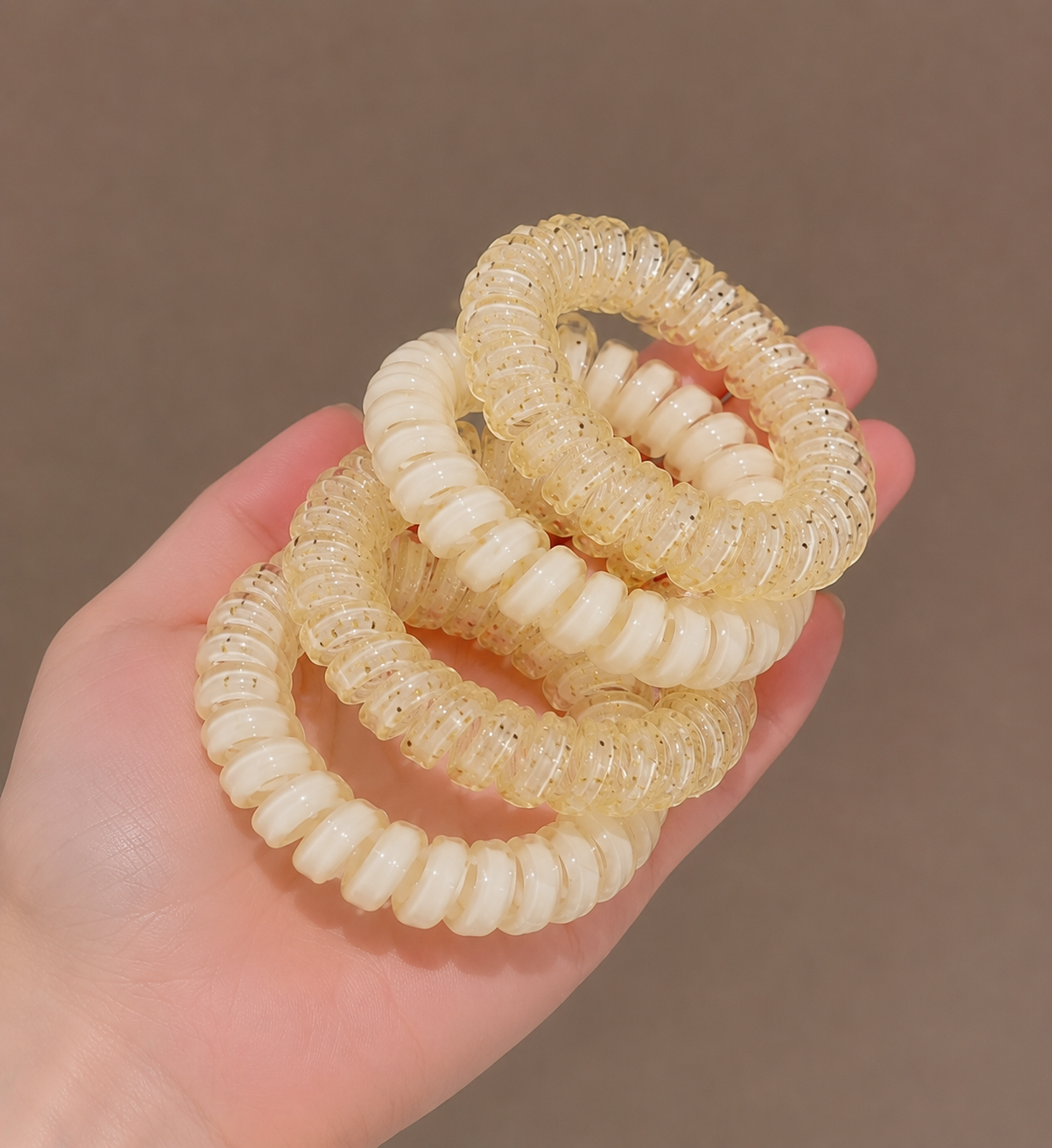Four translucent cream-colored spiral phone cord hair ties held in a person's hand against a solid taupe background.