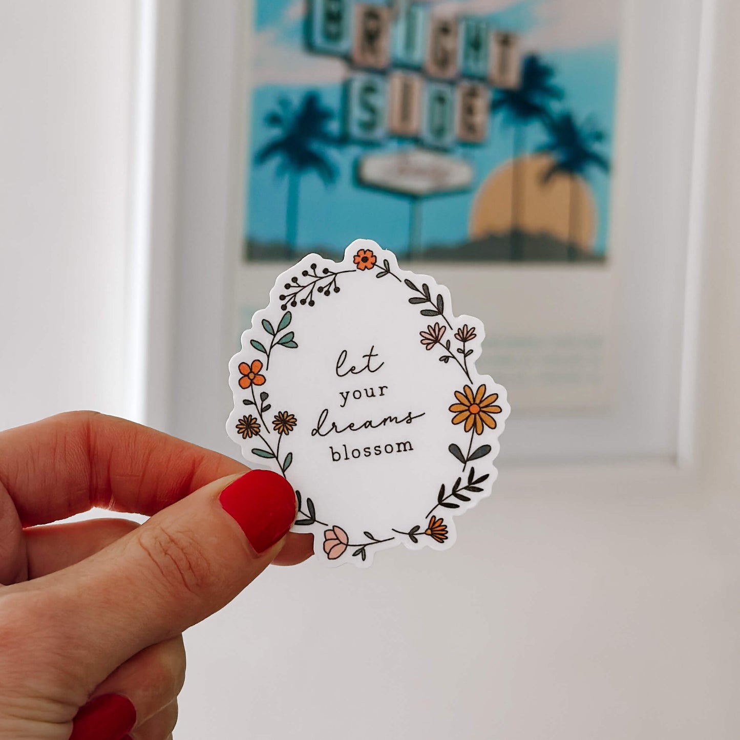 Hand holding vinyl sticker with flower illustration and the words 'et your dreams blossom' 