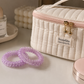 A quilted cream cosmetic bag with a pink zipper and gold hardware, featuring a 'Sozakka enjoy a better life' label. Two lavender spiral hair ties are in the foreground on a light beige surface, with a perfume bottle and a pink compact in the background.