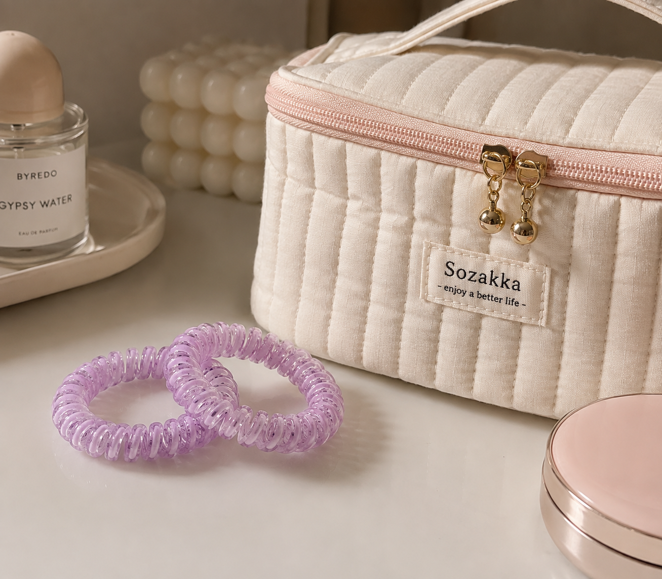 A quilted cream cosmetic bag with a pink zipper and gold hardware, featuring a 'Sozakka enjoy a better life' label. Two lavender spiral hair ties are in the foreground on a light beige surface, with a perfume bottle and a pink compact in the background.