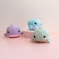 Three small crocheted whale amigurumi keychains in pastel colors, displayed on a pink surface with a light beige background. One is purple and pink, one is light blue, and one is mint green with a beige belly.