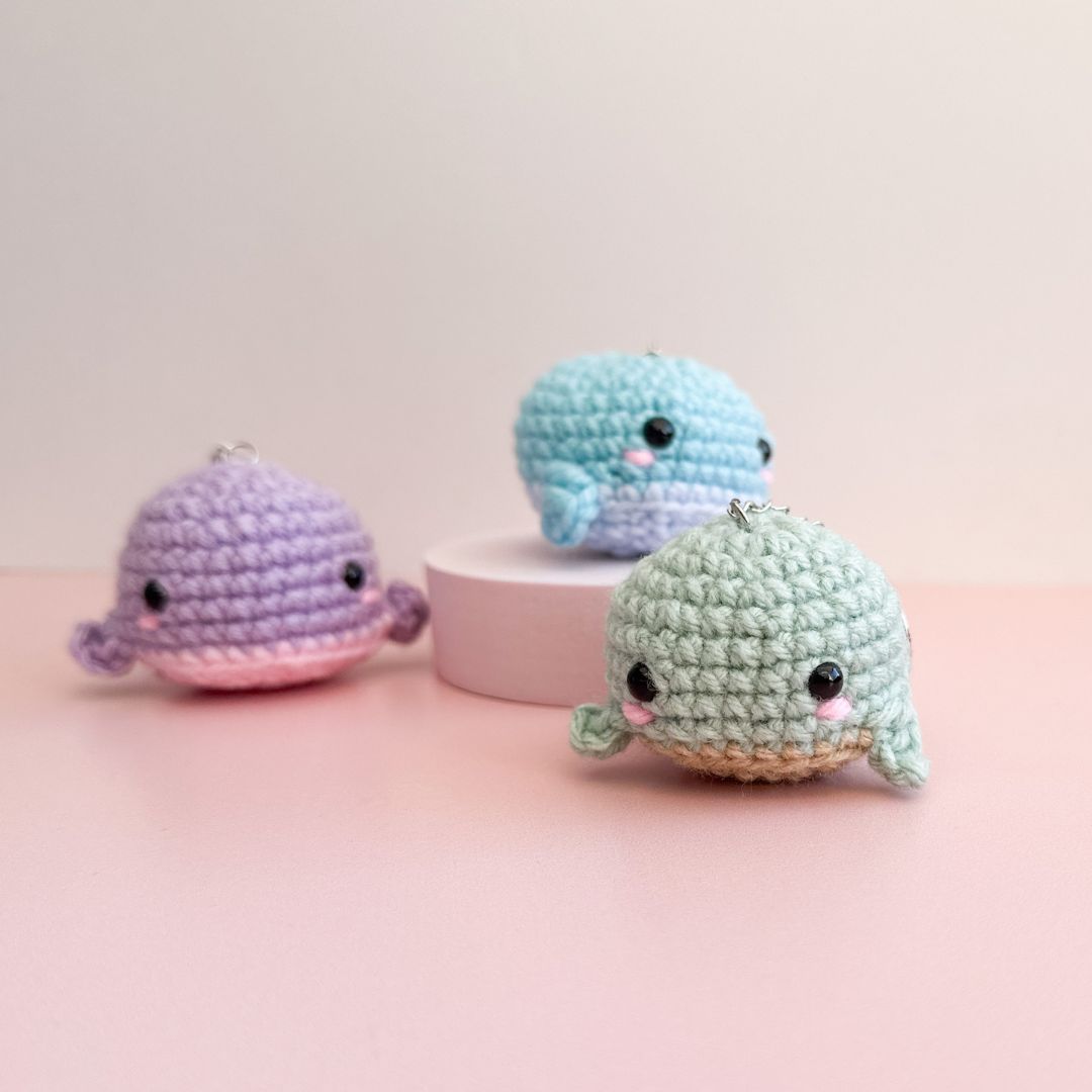 Three small crocheted whale amigurumi keychains in pastel colors, displayed on a pink surface with a light beige background. One is purple and pink, one is light blue, and one is mint green with a beige belly.